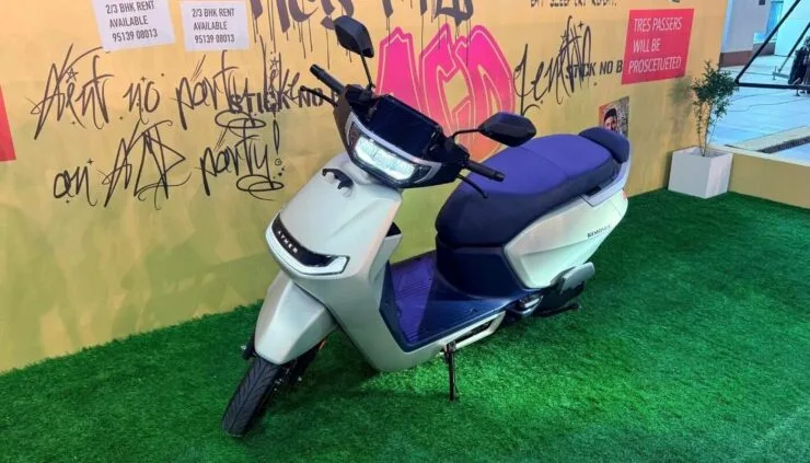 A front three-quarter view of the newly launched Ather Konarc electric scooter in silver and blue, parked on a green grass display with a graffiti wall background.