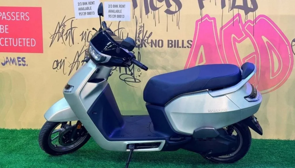 A full side profile view of the Ather Konarc electric scooter, showcasing its silver bodywork and blue seat on a green lawn at the launch.